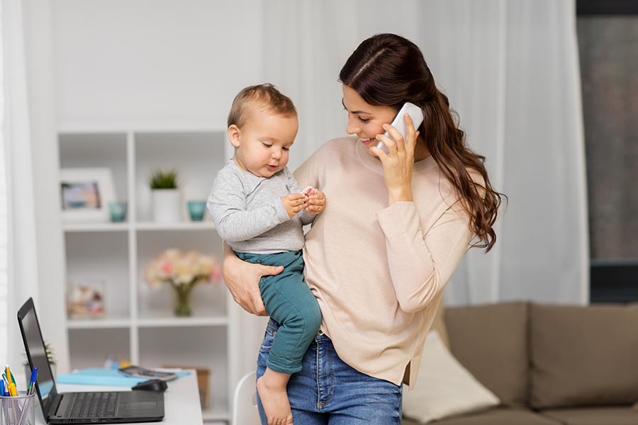 Contact Us - a Young Mom Makes a Call, Smiling and Balancing a Baby on Her Hip, Computer Open on a Nearby Desk in Their Beautiful Living Room