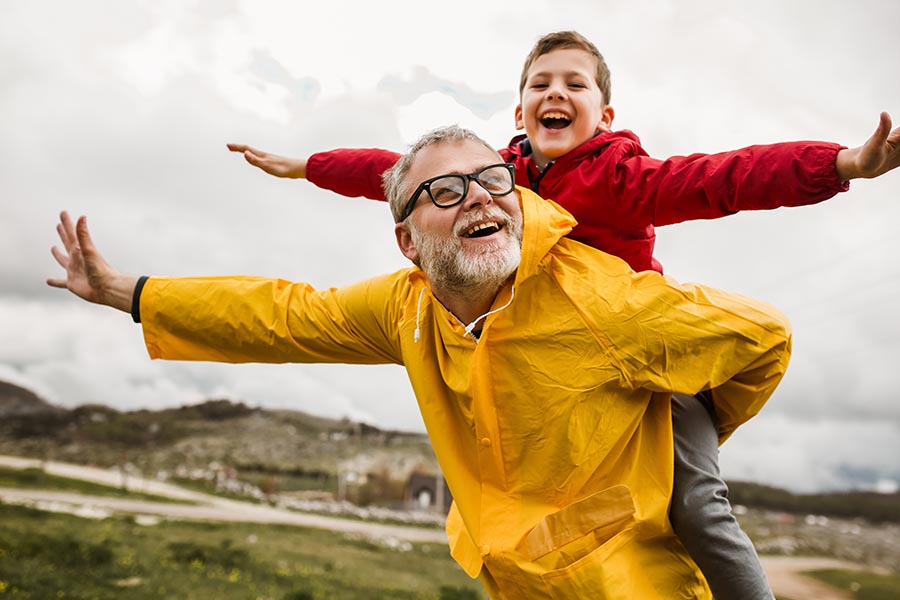 Personal Insurance - Young Boy Rides on His Grandfather's Back, Both Dressed in Rain Coats as They Walk Through a Green Landscape in the Rain